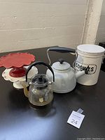 Photo showing light grey stove top kettle, two ceramic cake stands (red and white), white cookie tin marked 'COOKIES', and small glass teapot with diffuser inside on dark surface against light wall.