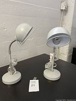Photo of two identical light grey desk lamps with curved necks and dome-shaped shades on round bases placed on a black table against a concrete wall.