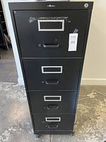 Front view of a black metal vertical filing cabinet with four drawers, each drawer having a label holder and a handle. The top two drawers are lockable with one key shown. Tape residue visible on the top drawer front. Cabinet on wheels.