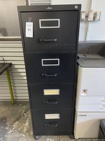 Photo of a black vertical filing cabinet with four drawers, each drawer has a handle and label holder. Lock cylinder visible on top drawer.
