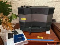 Front view of Bose system sitting on wood surface with power cord draped over top, remote controls and manual on left