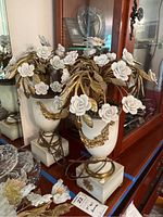 Pair of two identical ceramic flower table lamps with white urn-shaped ceramic bases, gold decorative motifs, topped with white ceramic roses and gold metal leaves. Arranged on wooden surface with blue tape line, visible cords for electrical connection.