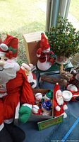 Overview of lot showing large stuffed Santa, tin, snowmen, wooden reindeer, decorative artificial holly tree, and assorted holiday figurines.