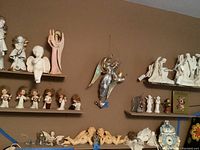 Wide view of shelves filled with various cherub and angel figurines in ceramic and cloth, a metal stained glass angel with lights hanging on wall, and a ceramic clock
