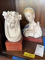 Two porcelain figurines by Cybis on wooden bases with one showing a female bust with bird, the other with floral wreath veil.