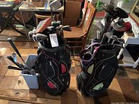Two black golf bags standing upright filled with golf clubs. The bags have red and purple/green accent panels. Plastic trash can with clubs nearby. Basement floor with wood planks showing.