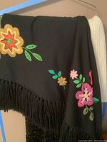 Black shawl with colorful floral embroidery and fringe hanging on a rack.