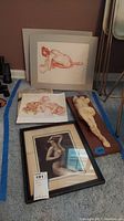 Photo showing a headless female nude sculpture mounted on wood, a framed nude black and white print or photograph, and two nude sketches on paper with warm red tones on floor.