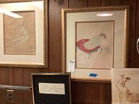 Three framed nude art pieces displayed on a wood panel wall, showing two nude sketches and one colored drawing.