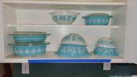 Three shelves containing Pyrex pieces: top Butterprint mixing bowl and Snowflake Blue covered dish; middle two Snowflake Blue covered casseroles; bottom three nested Snowflake Blue mixing bowls.