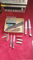 Full lot on red surface showing two Bowie knives with lucite handles, three Barlow knives, one red pocket knife, diamond whetstone and sheaths in box