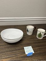 Four large white bowls stacked on a table with two ceramic Christmas mugs placed to the right.