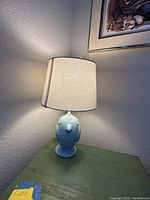 Blue ceramic bird-shaped lamp base with lit cream fabric lampshade on a green wooden table against a beige wall.