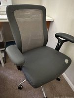 Front view of gray desk chair showing fabric seat with wear spot on right side and mesh backrest with padded armrests.