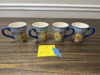 Four ceramic espresso cups with blue handles and floral designs placed in a row on wooden surface, showing one cup with a small chip on the brim.
