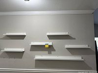 Six white floating ledge wall shelves mounted on a textured light beige wall in a staggered arrangement.