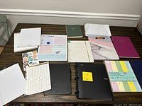 Photo showing multiple notebooks, notepads, a sheet of stickers, a small whiteboard sheet with marker, and color paper sheets arranged on a table.