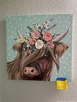 Close-up of square wall art depicting a cow with floral crown on light turquoise background with white spots, showing good condition.