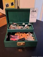 Green jewelry box open showing assorted jewelry inside: rings, earrings, brooches, some marked Ora and Trifar, plus some lipsticks below.