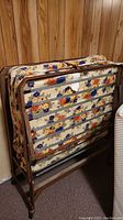 Folded single cot standing against wood panel wall with visible floral mattress.