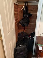 Photo showing a corner with five wheeled suitcases stacked and resting, a black handbag hanging, and a black backpack knapsack on wall hooks.