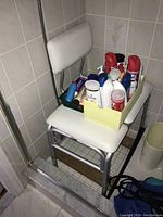 Shower chair with white padded seat and backrest with a yellow box of various grooming items on the seat inside a bathroom corner.