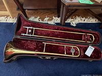 Full view of brass trombone inside a fitted hard protective case with red velvet lining, placed on blue carpet.