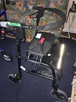Full view of the Nexus walker showing black padded seat, storage pouch underneath, foam padded handles, hand brake levers, and three-wheel design.