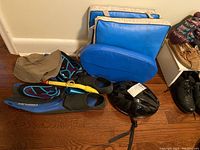 Photo showing two blue vinyl stadium cushions with beige straps stacked against the wall at the back, with water shoes, snorkel, flipper, hat and bike helmet on floor in front.