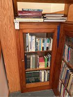 Overview of wooden cabinet filled with gardening and nature books, showing three shelves behind a glass door and additional books stacked on top.