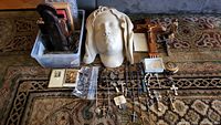Full lot overview showing crucifixes, wall hangings in plastic container, statue head, rosaries, prayer cards, and brass container.