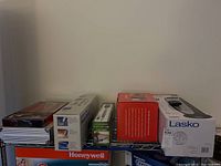 Photo of multiple boxed items on wire shelf including Lasko heater box, Brookstone speakers, and others