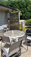 Outdoor patio setting with round glass top table, four metal frame chairs with mesh fabric seats and backs, and a metal wind chime hung nearby.