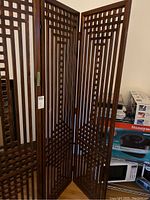 View of the front and side angles of the three-panel dark-stained wooden room divider with geometric lattice cutout pattern and metal hinges.