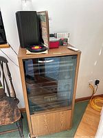 Wooden cabinet with glass front containing stereo components topped with vinyl records, speaker on top and remote control visible.
