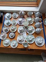 Photo of multiple tea cups, saucers, small tea pots, creamers, and lidded sugar bowls arranged on a wooden surface by a window.