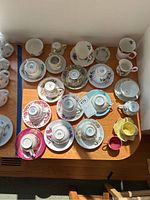 Overhead view of 14 assorted teacups and matching saucers including creamer, sugar bowl missing lid, and tea bag strainer.