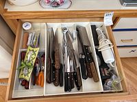 Top view of a kitchen drawer containing numerous knives with wooden and black handles, an electric carving cutter, sharpener, and other kitchen tools.