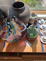 Overview of all lot items on wooden surface showing art glass vases, pottery jar, ceramic ginger jar, green glass pitcher in wicker basket, and porcelain teacup set