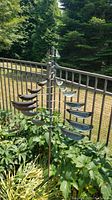 Outdoor vertical metal wind spinner with curved blades shown in garden bed surrounded by greenery and railing in background.