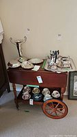 Full view of teacart showing double drop leaves, top wooden surface, drawer, and wheels. Vintage teacups are on bottom shelf.