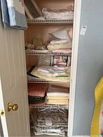 Photo of linen closet shelves holding various folded bedspreads, pillow shams, and assorted linens in neutral, floral, and pastel colors.