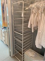 Tall white metal wire basket shelving unit with nine baskets in varying widths, the unit stands beside hanging white clothes.