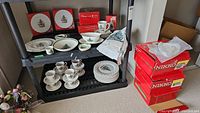 Wide shot showing dinner plates, cups and saucers, gravy boat, coffee mugs, small plates, arranged on black shelf next to stacks of red Nikko original boxes