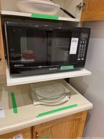 Front view of black Panasonic inverter microwave on white shelf with three clear glass turntable plates underneath