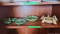 Wide shot of three green cabbage leaf serving plates and beige goose flower frog vase on wooden shelf.