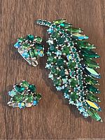 Close-up of Sherman brooch and two clip-on earrings showcasing green and blue rhinestones arranged in leaf and floral patterns on a wooden surface.