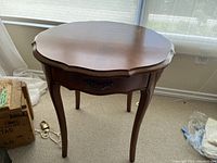 Front angle showing the wooden side table with scalloped edge and drawer with metal pull handle. Light brown finish visible.