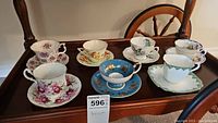 Image shows all six teacups with matching saucers, arranged on a wooden surface with a MaxSold tag 596 visible