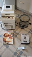 Photo showing both the Hitachi bread maker with cookbook and the HomeImage 2-tier steam cooker with manual, placed on a patterned floor.